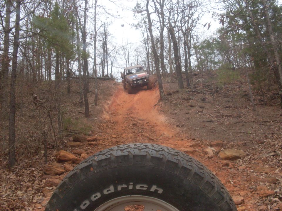 View from inside a vehicle looking at another off-road vehicle climbing a steep, red dirt trail in a wooded area
