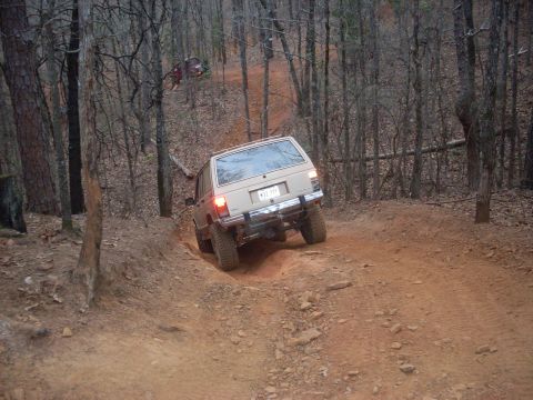 1996 Ford Bronco - A tan Jeep Cherokee XJ navigates a steep, muddy off-road trail surrounded by trees.