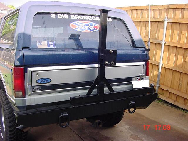 Rear of blue and silver Ford Bronco with custom black bumper and swing-out tire carrier, parked by a wood fence