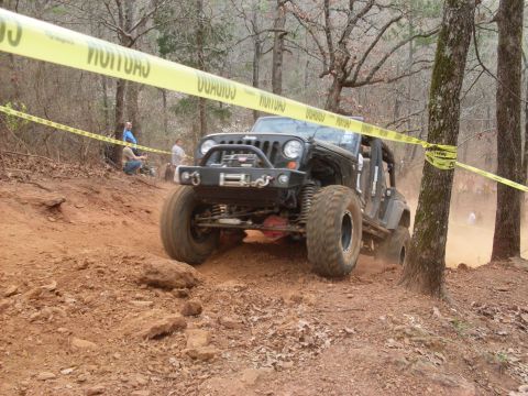 1996 Ford Bronco - A black Jeep Wrangler tackles a challenging off-road trail, marked by caution tape, during an event.