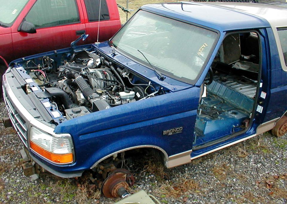 Blue Ford Bronco being stripped, hood off, door and front wheel removed, in a salvage yard