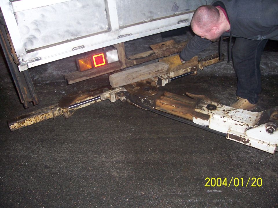Trailer hitch mechanism with a man inspecting it. Rusty metal and wood supports are visible