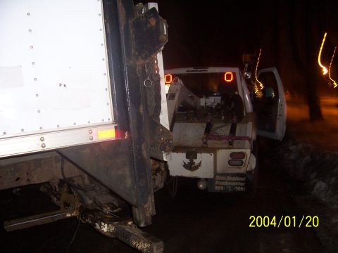 1990 Ford Bronco - A tow truck is parked next to a white box truck at night, with equipment visible in the tow truck's bed.