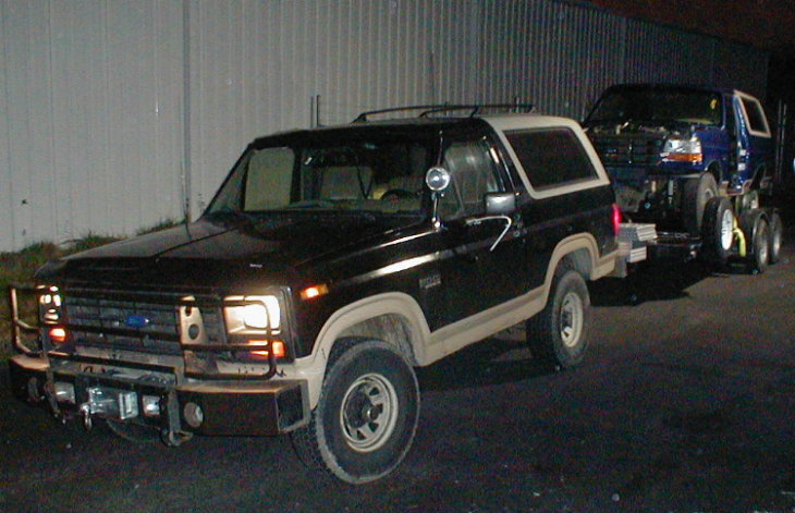 Black and tan Ford Bronco at night towing a blue pickup on a trailer beside a metal building