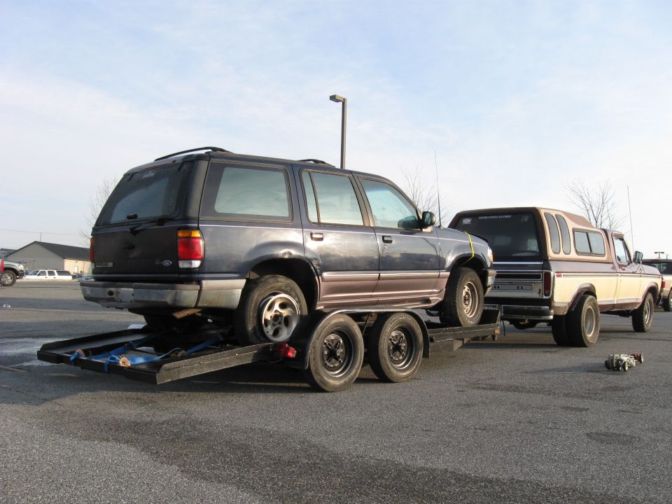Rear view of a blue Ford Explorer on a trailer towed by a two-tone pickup