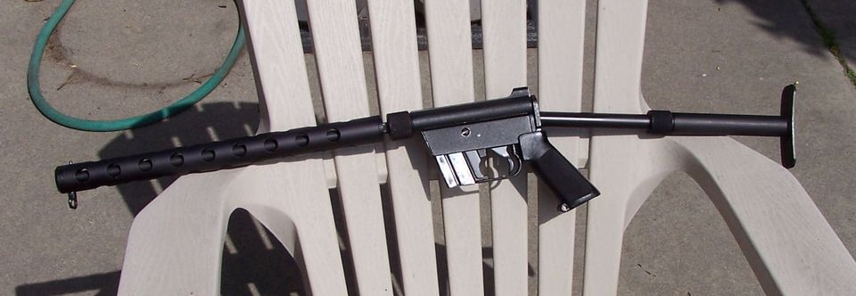 Black submachine gun resting on a white plastic chair, with a green hose in the background