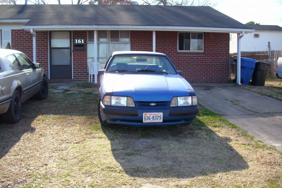 Blue Fox-body Ford Mustang sedan parked on a lawn in front of a brick house