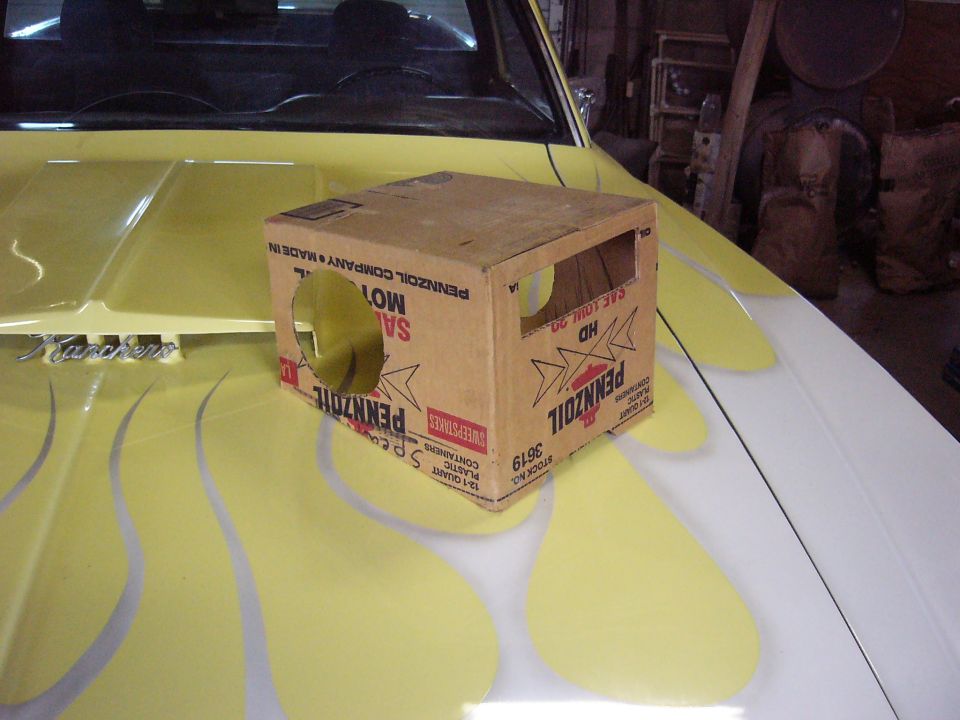 Cardboard box with Pennzoil logos sits on the hood of a yellow car with flame decals. The car has 'Ranchero' badging
