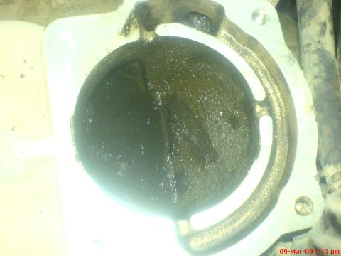 1992 Ford Crown Victoria - Overhead flash close-up into a throttle body bore, showing the closed throttle plate and bore walls glazed with wet, oily black carbon buil…