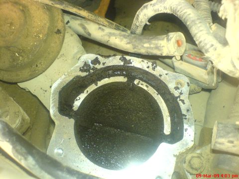 1992 Ford Crown Victoria - Angled close-up of an engine-mounted throttle body with the inlet duct off, showing the closed throttle plate and bore blanketed in thick b…