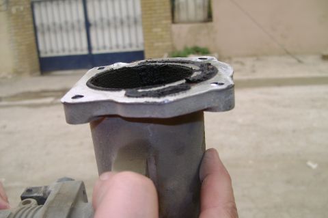 1992 Ford Crown Victoria - A hand holds up an aluminum throttle-body-style spacer or intake elbow outdoors, tipping it to show the mating flange packed with thick bla…