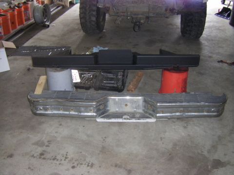 1989 Ford Bronco - Two bumpers are displayed in front of a truck's rear axle and tires in a garage setting.