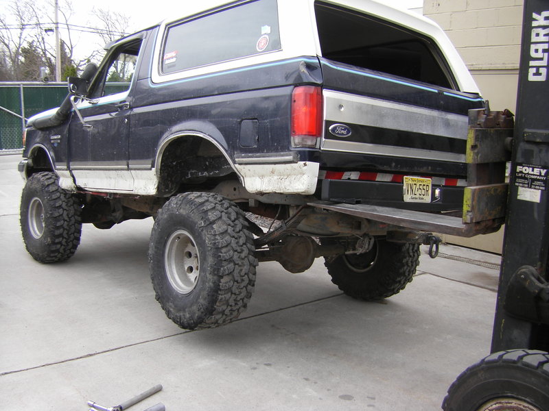Lifted blue and white Ford Bronco on mud tires with rear bumper raised by a forklift