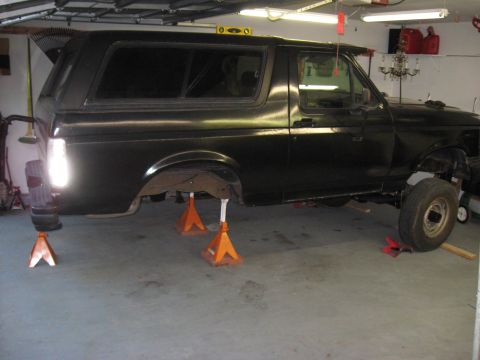 1991 Ford Bronco - A black Ford Bronco is elevated on jack stands in a garage, showing its undercarriage and missing rear wheel.