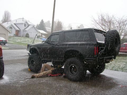 1996 Ford Bronco - A person works on the underside of a lifted black Ford Bronco in a driveway as snow falls.