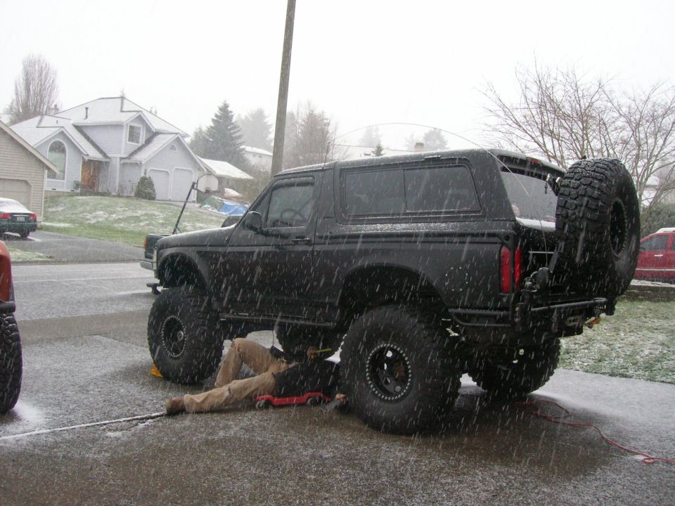 Black 1996 Ford Bronco lifted with large tires, person working underneath during snowfall