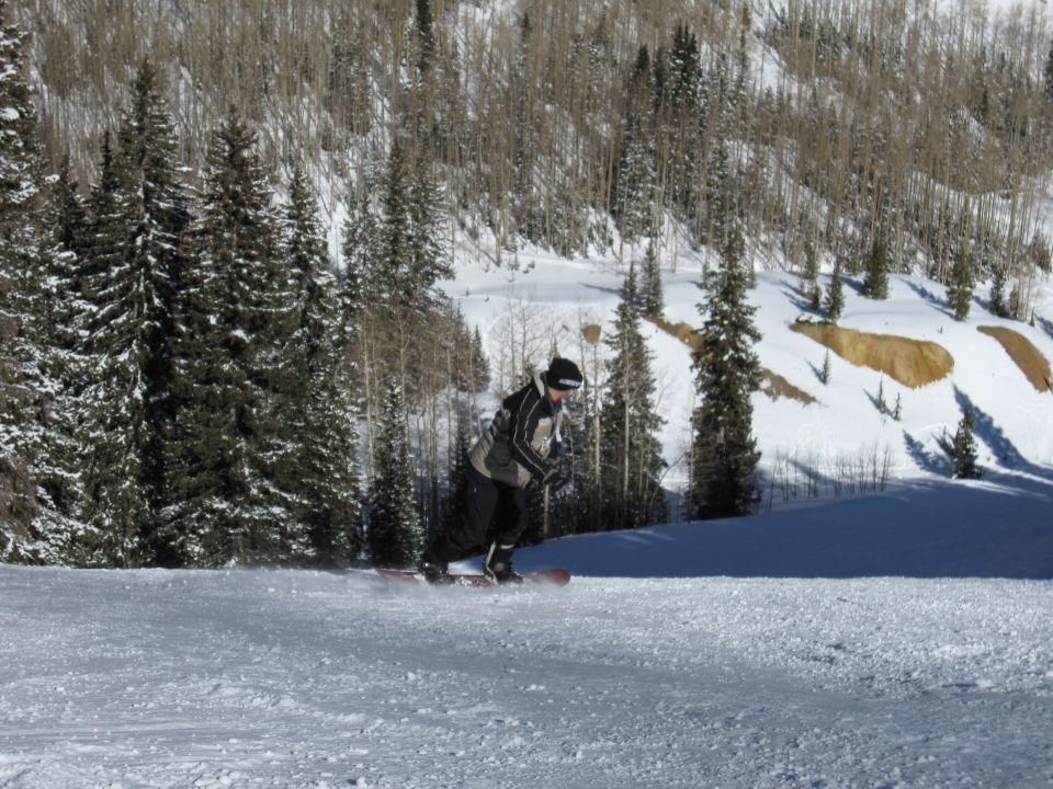 Person snowboarding down a snowy mountain with pine trees and bare trees in the background. Watermark says SUPERMOTORS.NET