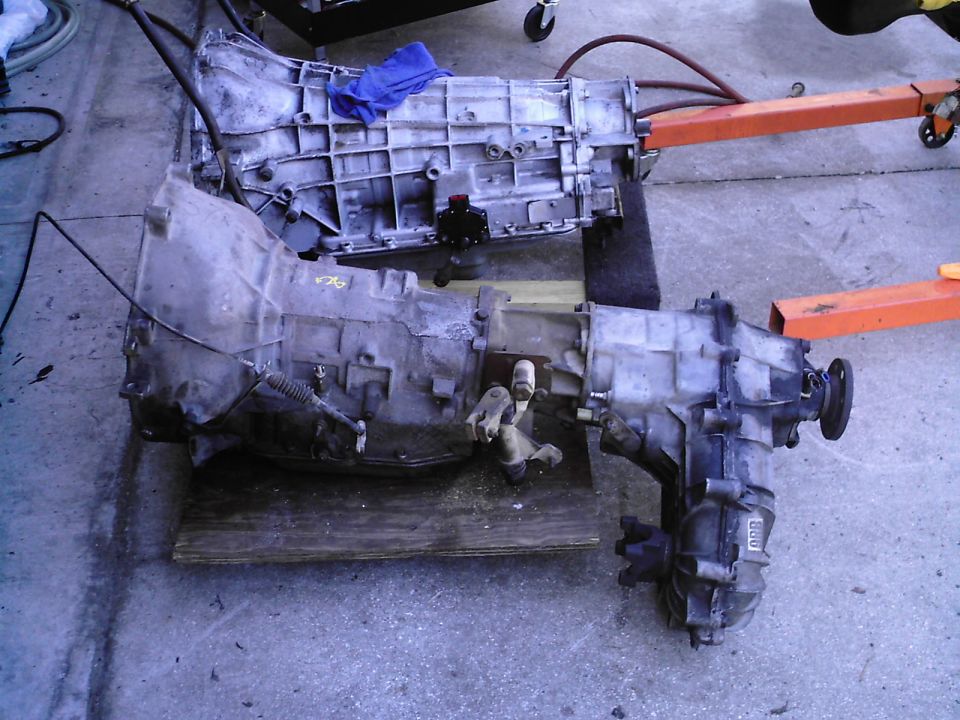 Two automatic transmissions on plywood on concrete, one with transfer case attached, orange hoist legs nearby