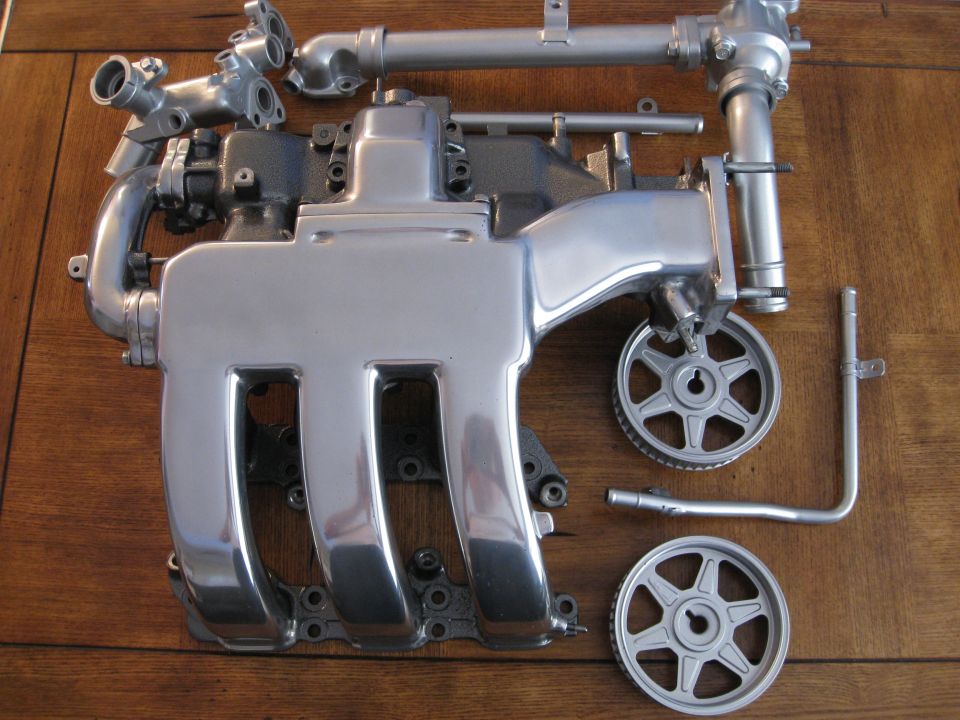 Engine intake manifold with pipes and two pulleys laid out on a wooden surface. Supermotors.net logo visible