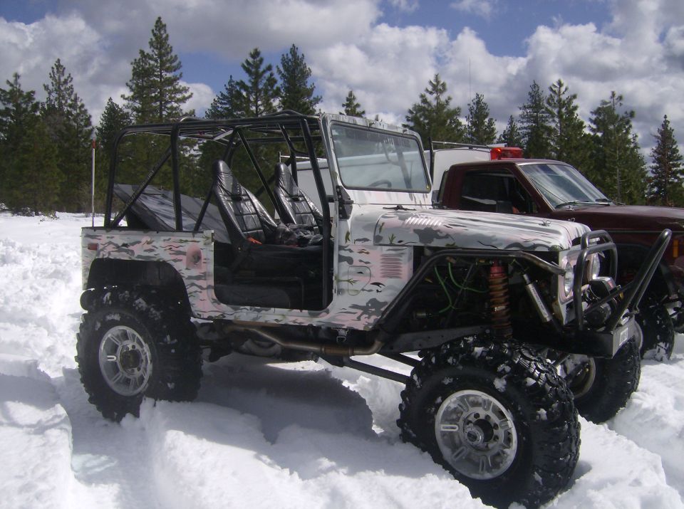 Snow-camo open-top 4x4 with roll cage and tube bumper parked in deep snow by pines