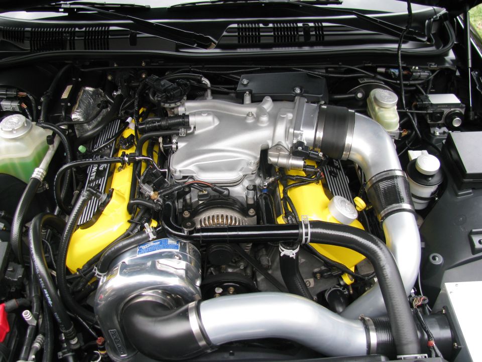 Engine bay with a polished supercharger, yellow valve covers, and silver intake piping. Supermotors.net logo visible