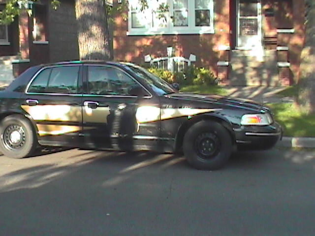 Black Ford Crown Victoria sedan with black steel wheels, side view, parked on a shaded residential street