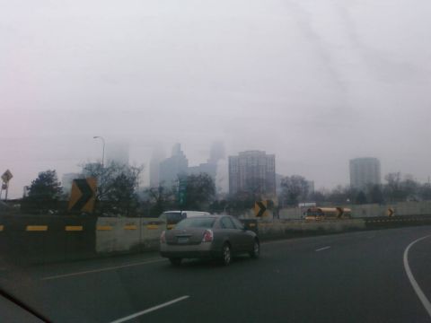 1985 Ford LTD Crown Victoria - A gray sedan drives on a highway with a foggy city skyline visible in the distance.