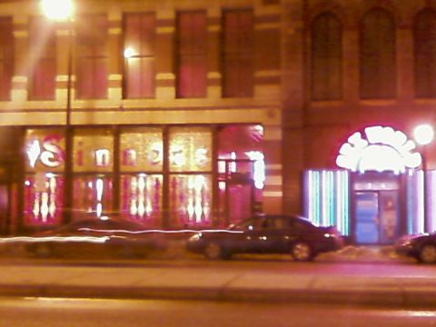 1985 Ford LTD Crown Victoria - A dark sedan is parked on the street in front of a building with neon lights.