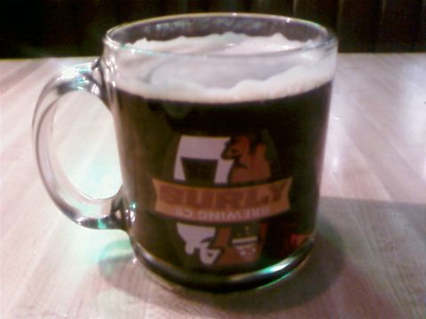 1985 Ford LTD Crown Victoria - A glass mug of dark beer with a prominent Surly Brewing Co. logo is displayed on a wooden surface.