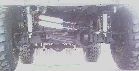 1986 Ford Bronco - Underside view of a solid front axle on a lifted 4x4, showing the differential housing, coil springs, dual steering stabilizers with red ac…
