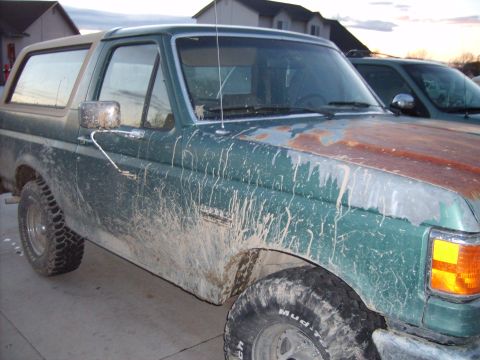 1989 Ford Bronco - Mud-splattered green 1989 Ford Bronco seen from the front three-quarter angle, with another vehicle visible behind it.