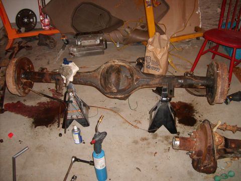 1989 Ford Bronco - A heavily rusted rear axle assembly is supported by jack stands in a garage setting, surrounded by tools and a propane torch.