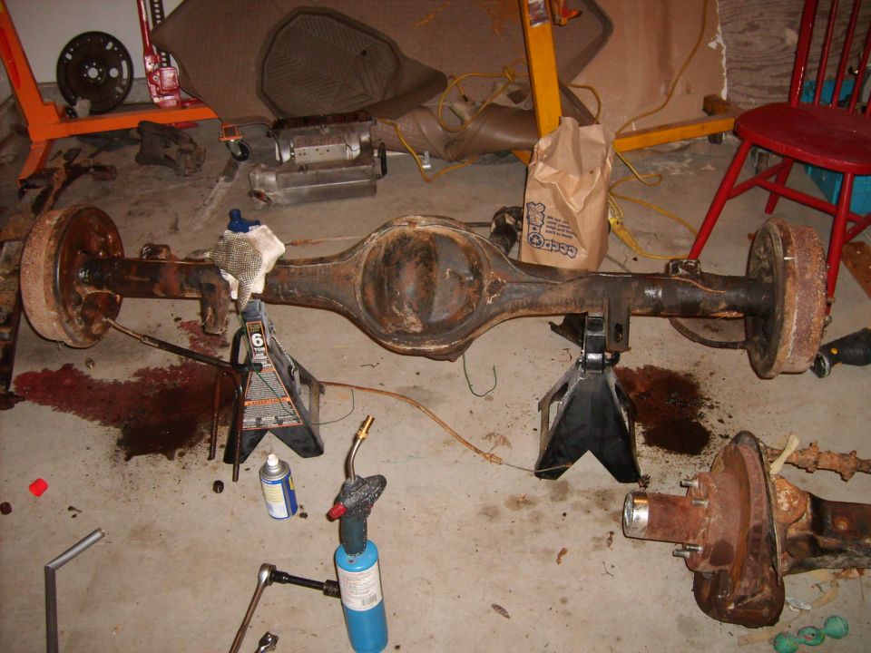 Rusty rear axle assembly on jack stands with tools and a propane torch nearby