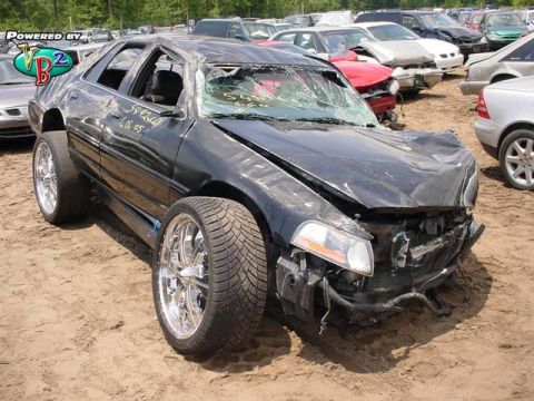 1993 Ford Crown Victoria - A heavily damaged black 1993 Ford Crown Victoria sits in a junkyard with other wrecked vehicles.