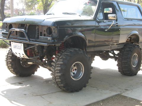 1987 Ford Bronco - Modified black Ford Bronco with large tires and a custom front bumper sits on pavement.