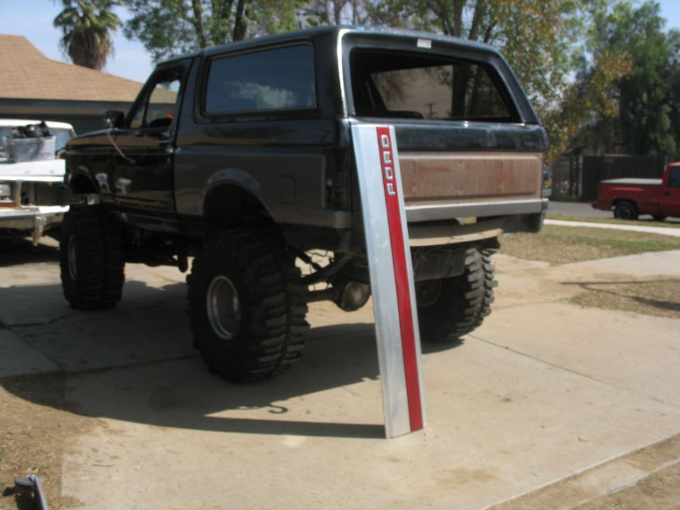 Lifted black Ford Bronco with large tires, parked outside. A silver and red Ford trim piece is leaning against the rear