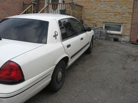 2003 Ford Crown Victoria - A white sedan is parked in front of a brick building with a driveway.