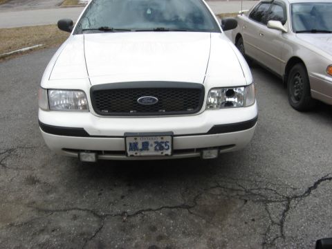 2003 Ford Crown Victoria - Front view of a white 2003 Ford Crown Victoria, showing its grille and headlights. Another car is parked to the right.