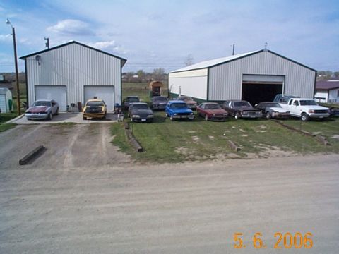 1984 Ford Mustang - A collection of vehicles are parked outside two large garages on a cloudy day.