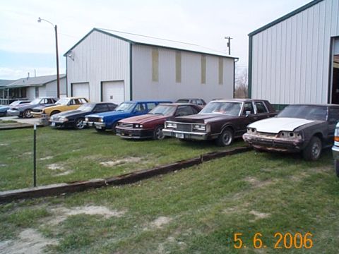 1984 Ford Mustang - A row of cars is parked outside buildings, including a dark coupe with a white hood at the front.