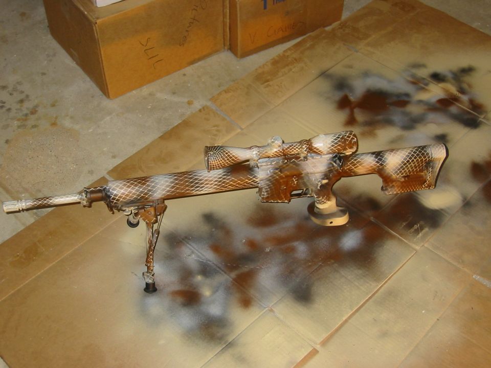 Sniper rifle with camouflage paint job, resting on cardboard
