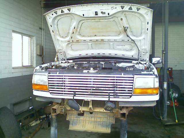 White Ford Bronco front end with hood up, bumper removed, on jack stands in garage