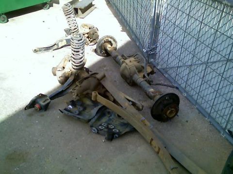 1996 Ford Bronco - Various vehicle suspension components and axles are laid out on a concrete surface, with a chain-link fence visible behind them.