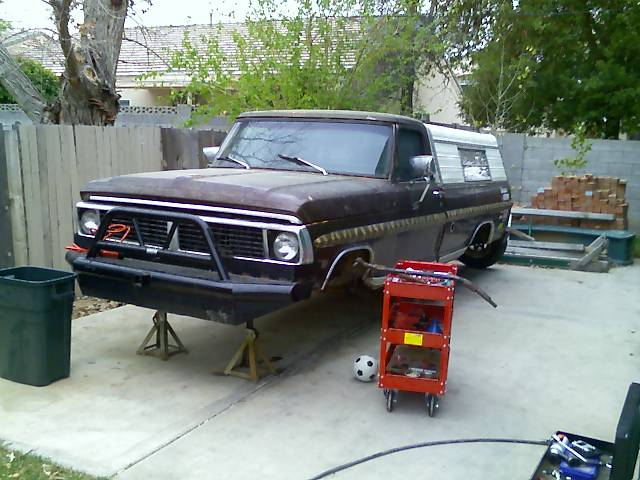 Faded brown 1970s Ford pickup with camper shell on jack stands in a backyard driveway