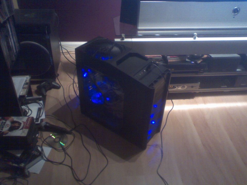 Black computer tower with blue LEDs on a wooden floor with wires and gaming accessories.