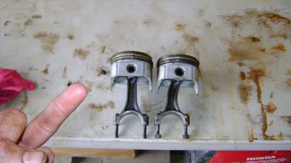 Two engine pistons on connecting rods on a workbench with a finger pointing at them