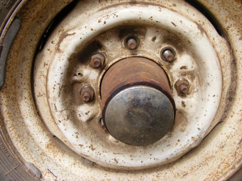 1985 Ford Bronco - A close-up, slightly angled view of a rusty, white wheel hub with five lug nuts and a metal center cap. The tire is visible on the left sid…