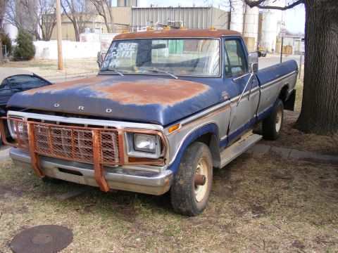1985 Ford Bronco - A rusty blue and silver Ford pickup truck is parked on a dirt and grass lot next to a large tree. The truck has a large orange metal grille…