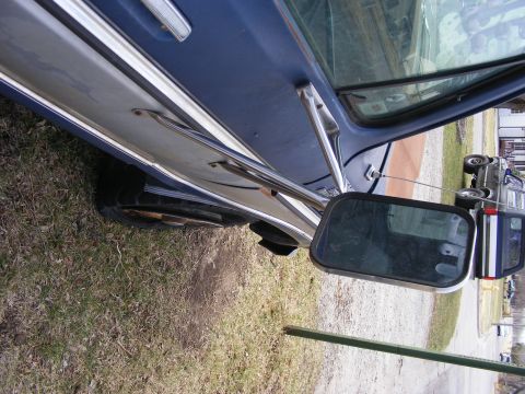 1985 Ford Bronco - The side of a blue and silver vehicle is shown, with a large side mirror reflecting the surroundings. The vehicle appears to be parked on a…