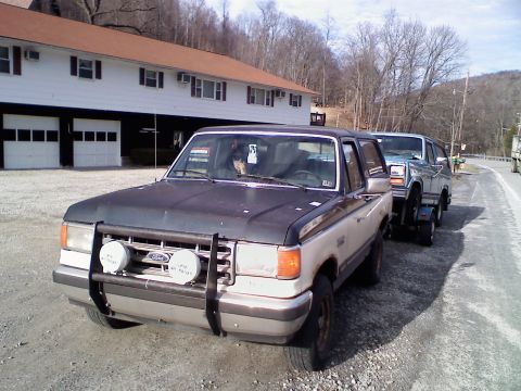 1988 Ford Bronco - A two-tone Ford Bronco, with a dark gray hood and a tan lower body, is parked in front of a building with two garage doors. A second, light…